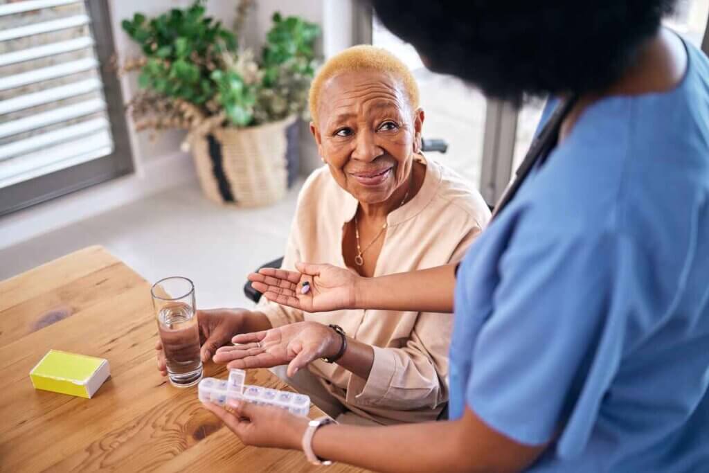 Nurse, smile and giving old woman pills in home with water for healthcare, wellness and support.