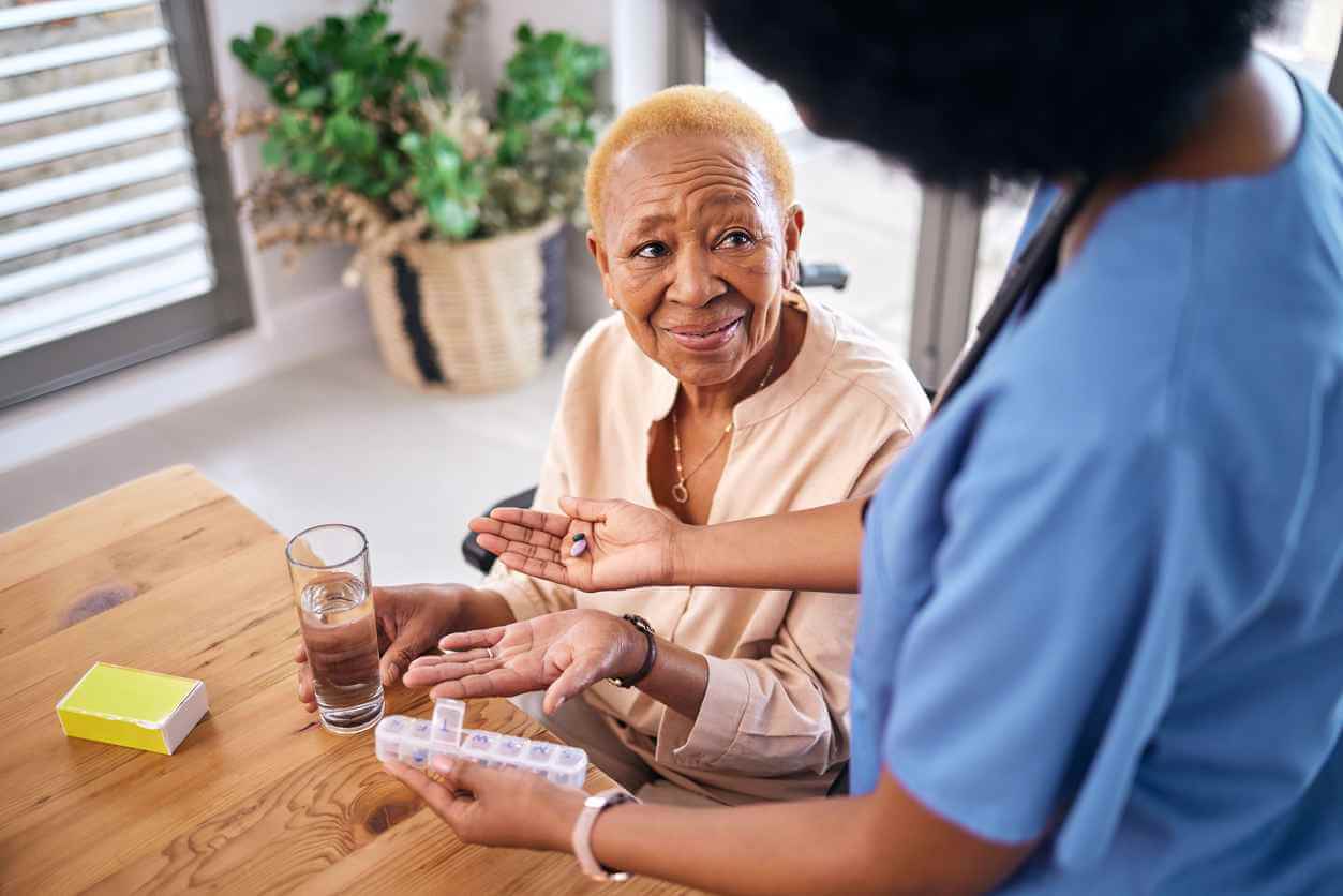 Nurse, smile and giving old woman pills in home with water for healthcare, wellness and support.