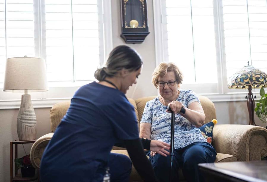 An elderly woman has a home visit with her nurse who is helping her around the house.