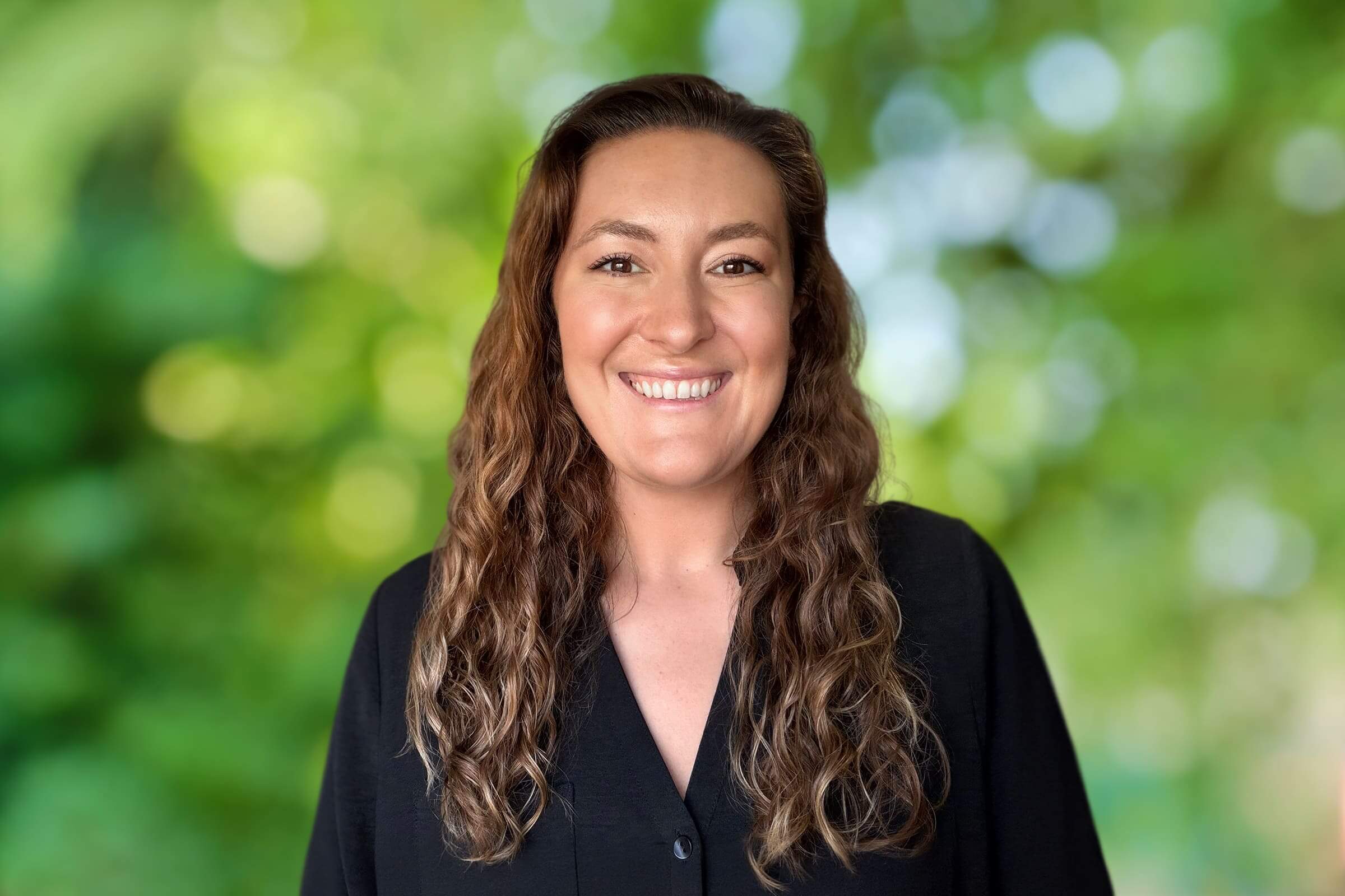 Smiling woman with wavy brown hair wearing a black top, against a blurred green outdoor background.