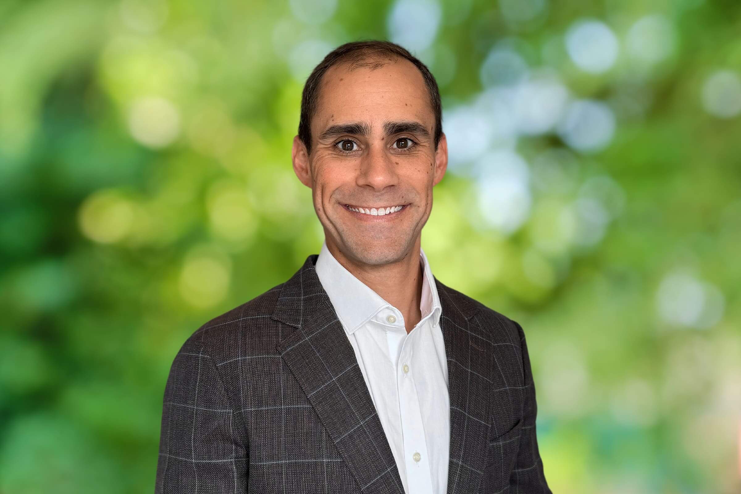 Smiling man in a dark checkered suit and white shirt, posing outdoors with a blurred green leafy background.