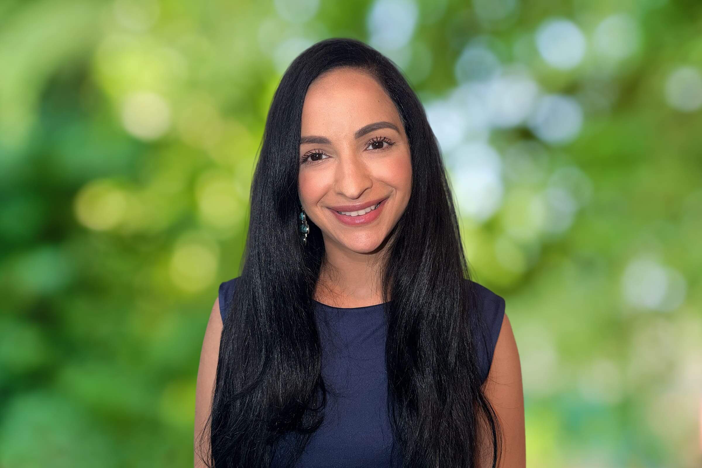 Close-up portrait of a smiling woman with long black hair wearing a navy top and a green, blurred outdoor background.