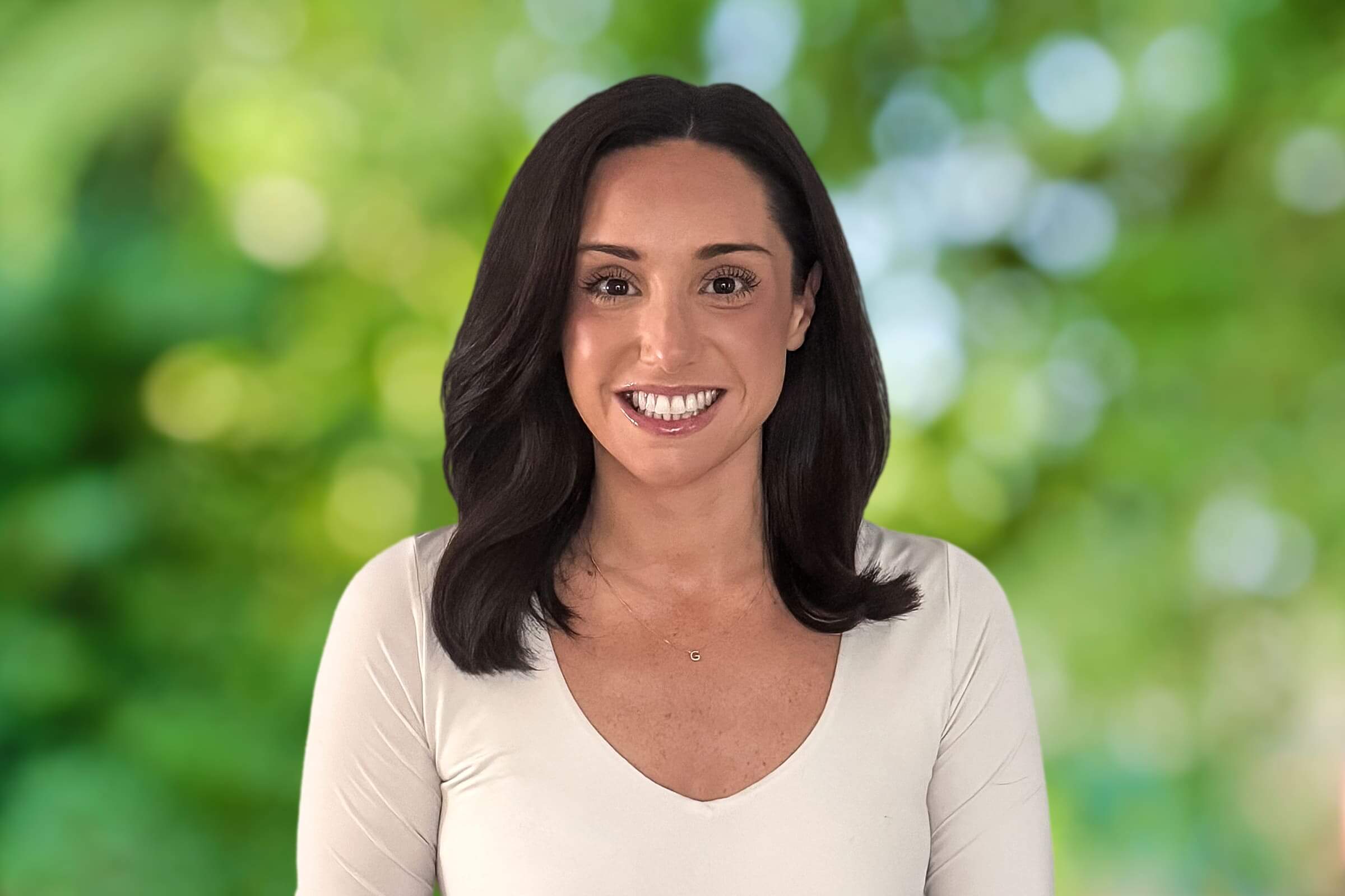 Smiling woman with shoulder-length dark hair in a light top, outdoors with a blurred green background.