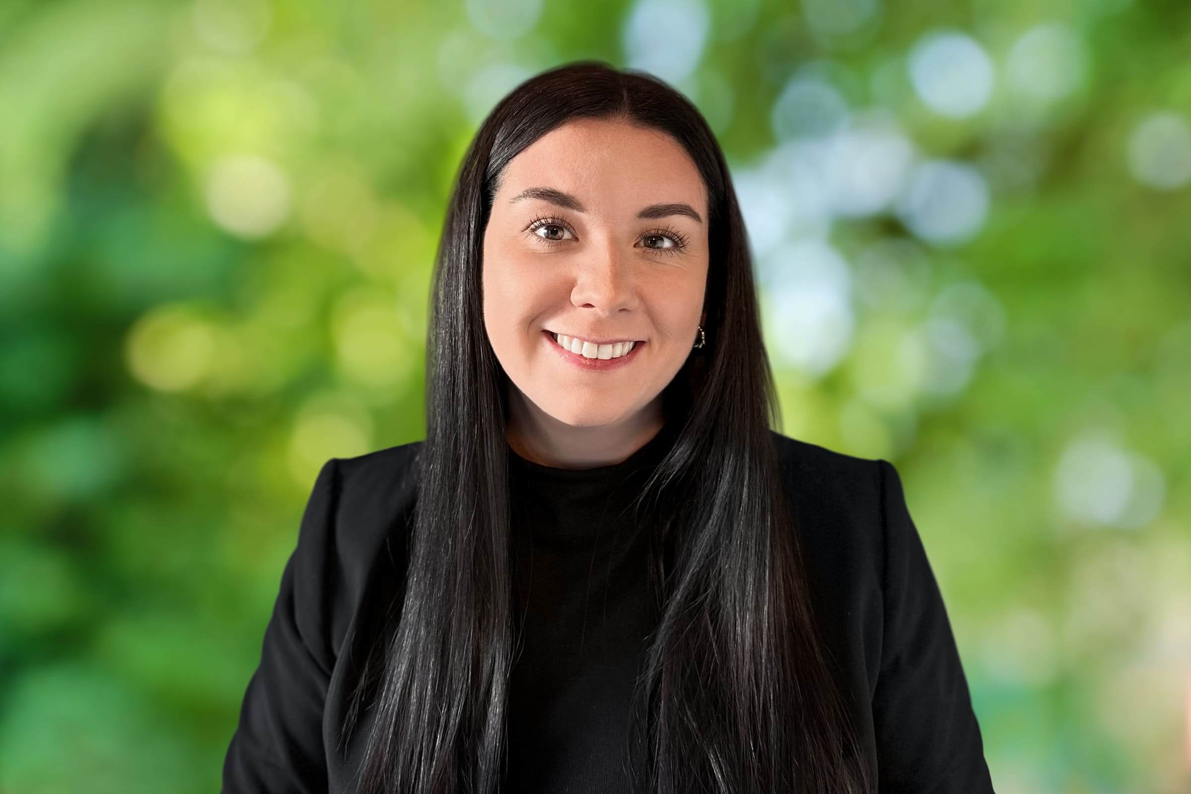 Portrait of a smiling woman with long black hair wearing a black top, outdoors with a green, leafy background.